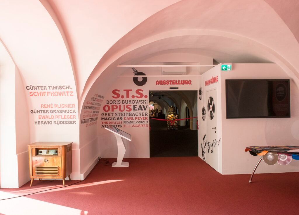 Ein Innenraum mit rosa Wänden und rotem Teppich, der eine Ausstellung von musikbezogenen Kunstwerken zeigt. Ein hölzerner Schrank mit einem Radio steht links, und ein weißer Ständer mit einem Bildschirm befindet sich in der Mitte. Die Wände sind mit Namen und Musiknoten verziert.