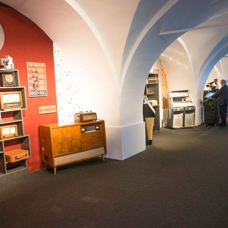 Ein Raum mit roter Wand und gewölbter Decke, der alte Radios und ein Poster mit dem Titel 'The Birth of Rock n' Roll' zeigt. Im Hintergrund stehen zwei Männer, einer mit einer Kamera.