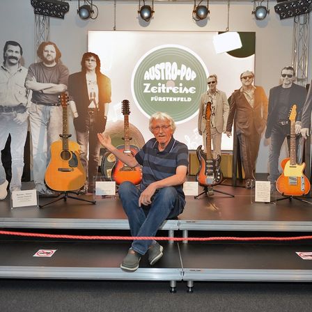 Ein Mann sitzt auf einer Bühne vor einer Kulisse mit einer Gruppe von Musikern und Gitarren. Er trägt eine Brille und ein gestreiftes Shirt. Hinter ihm befinden sich Ausschnitte von Musikern mit Gitarren und ein großer Kreis mit Text.