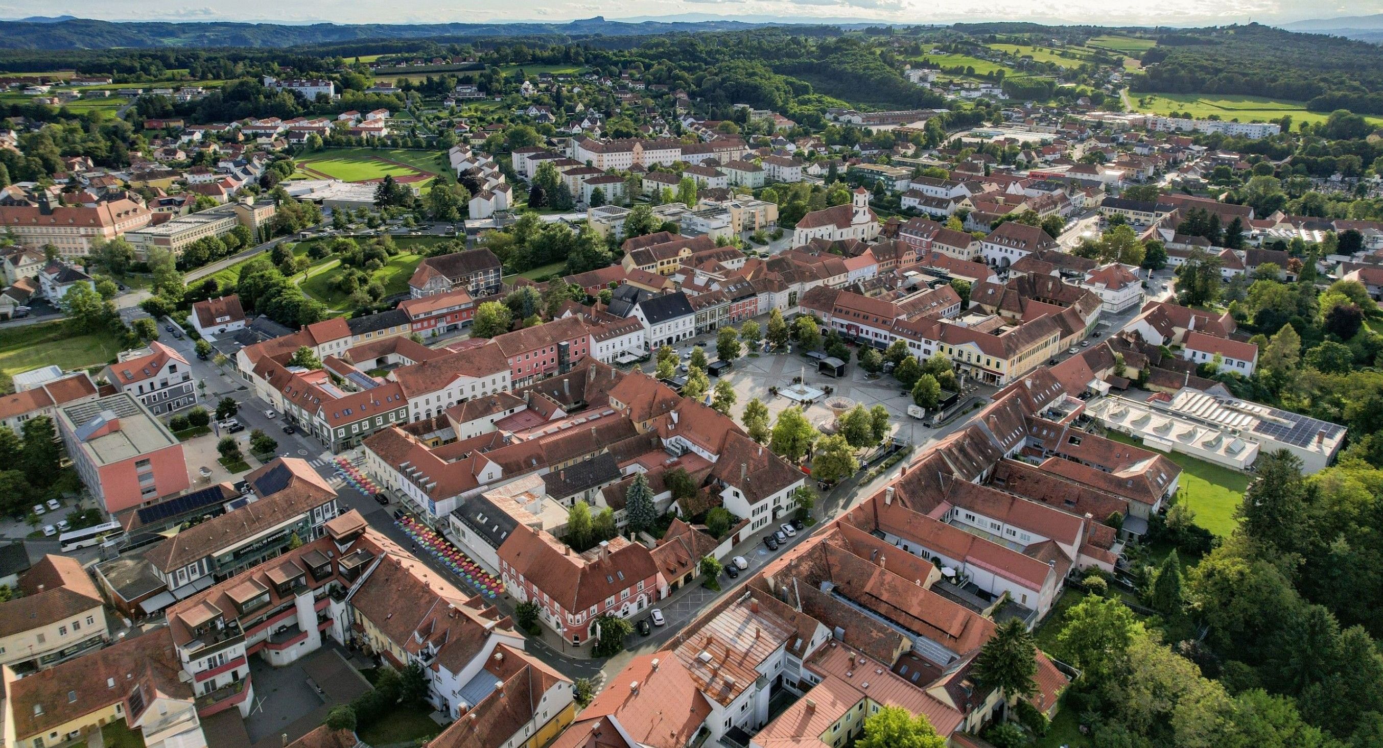Luftaufnahme einer Stadt mit vielen Gebäuden, roten Dächern und grünen Bäumen. Die Stadt ist von Hügeln und einem klaren blauen Himmel umgeben.