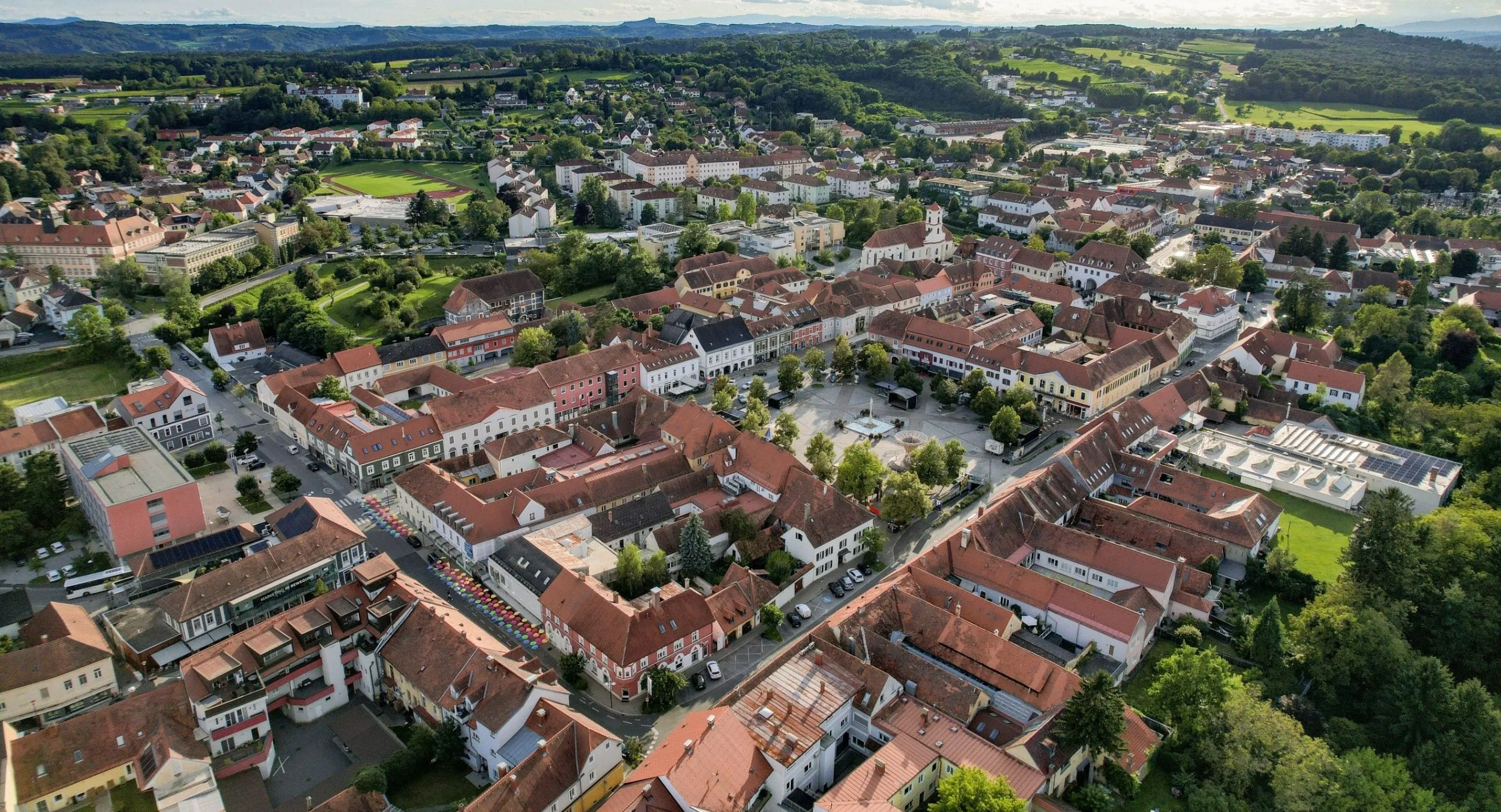 Luftaufnahme einer Stadt mit vielen Gebäuden, roten Dächern und grünen Bäumen. Die Stadt ist von Hügeln und einem klaren blauen Himmel umgeben.
