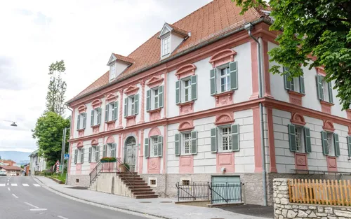 Ein rosa und weißes Gebäude mit vielen Fenstern und Fensterläden. Eine Treppe führt zum Eingang. Das Haus hat ein braunes Dach und befindet sich an einer Straße.