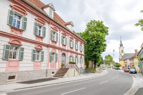 Ein rosa und weißes Gebäude mit mehreren Fenstern und grünen Fensterläden. Eine Treppe mit Geländer führt zum Eingang. Ein Baum steht neben dem Gebäude. Die Straße ist leer mit einer weißen Linie.