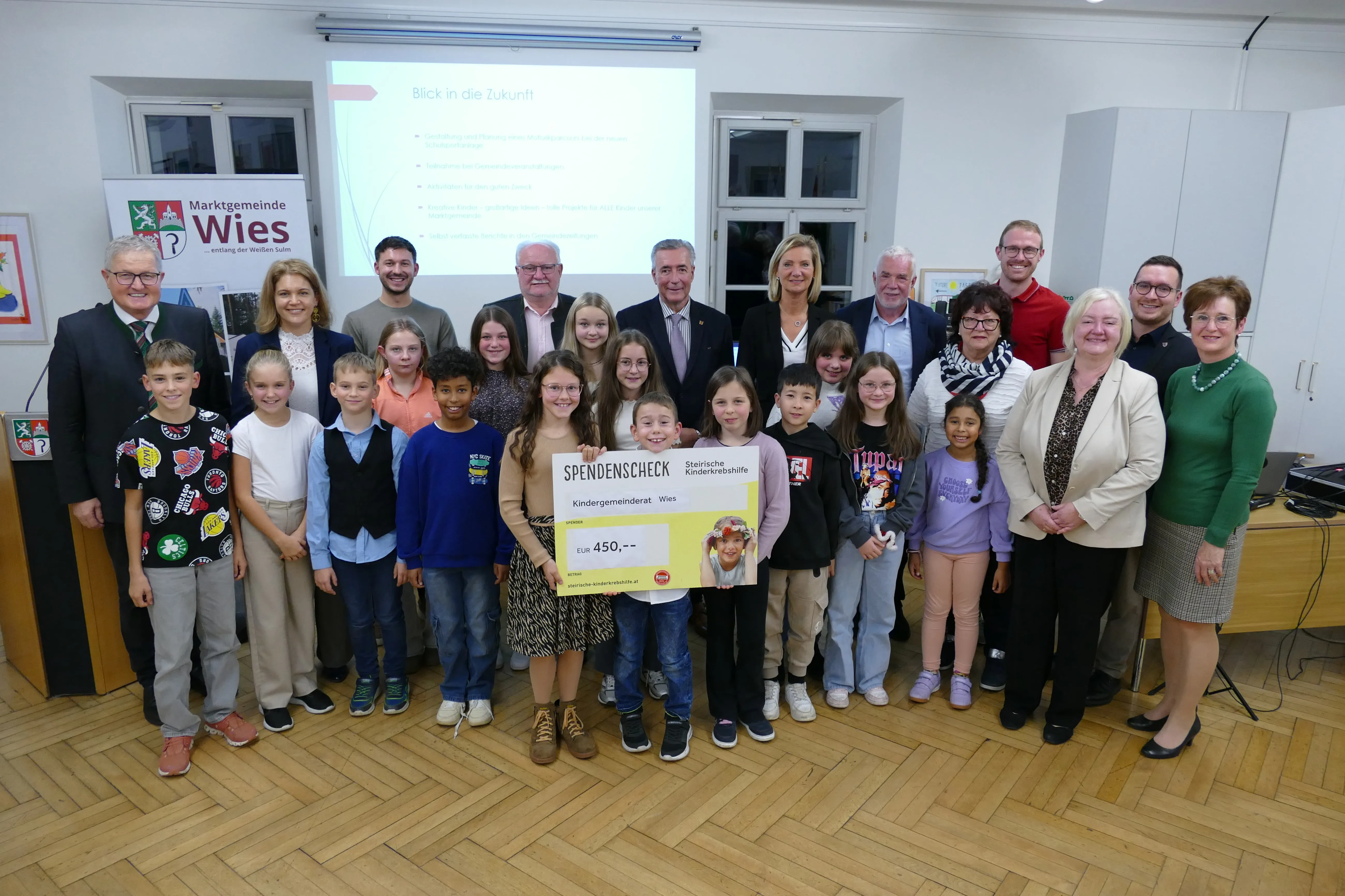 Eine Gruppe von Erwachsenen und Kindern posiert für ein Foto in einem Raum mit einer Projektionsleinwand, die Text anzeigt. Ein Kind hält einen Scheck mit der Aufschrift 'SPENDENSCHECK' und einem Bild eines Kindes darauf.