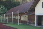 Ein eingezäuntes Gebäude mit braunem Dach und Metallzaun steht auf einem Rasenfeld mit einer roten Laufbahn. Bäume und Grün umgeben das Gebiet.