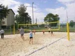 Eine Gruppe von Menschen spielt Volleyball auf einem sandigen Spielfeld mit einem gelben Netz und gelben Stangen. Ein Gebäude und Bäume sind im Hintergrund.