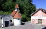 Eine kleine Kapelle mit einem roten Dach und einem Glockenturm befindet sich zwischen zwei Gebäuden mit Garagen. Ein Gebäude ist rosa mit weißer Umrandung und das andere ist weiß mit einem roten Dach. Bäume sind im Hintergrund.