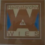 Ein braunes und beiges Logo mit dem Text 'Heimatarchiv' oben und 'WIES' unten mit einem blauen Dreieck in der Mitte.