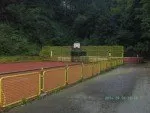 Ein Tennisplatz im Freien mit gelben und braunen Zäunen, umgeben von Bäumen und Grün. Der Platz scheint leer zu sein.