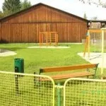 Ein Freiluftspielplatz mit einem Holzschuppen, einer Bank und Spielgeräten. Der Bereich ist mit einem gelb-grünen Netz eingezäunt, und in der Nähe befindet sich ein Mülleimer.