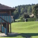 Ein hölzerner Pavillon steht auf einem Hügel mit einer farbenfrohen Wendeltreppe. Der Hügel ist üppig mit grünem Gras und Bäumen bedeckt, und ein Haus ist in der Ferne zu sehen.