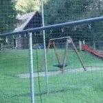 Ein Spielplatz auf einer Rasenfläche mit einer Schaukel, einer Rutsche und einem Holzhaus im Hintergrund.