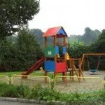Ein Spielplatz mit einem bunten Rutschen, Schaukeln und einer grünen Schaukelsitz, umgeben von Pflanzen und Bäumen.