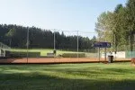 Ein Tennisplatz im Freien mit Zaun und Mülleimer. Der Platz ist von grünem Rasen und hohen Bäumen umgeben. Der Himmel ist klar und blau.