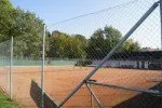 Ein Outdoor-Tennisplatz mit einem Maschendrahtzaun und einem Netz. Der Platz ist leer, und es gibt Bäume im Hintergrund.
