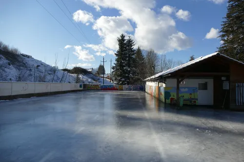 Eine leere Eisbahn mit bunten Graffiti-Wänden und einem verschneiten Hügel dahinter. Bäume umgeben die Bahn und ein kleines Gebäude ist rechts zu sehen.
