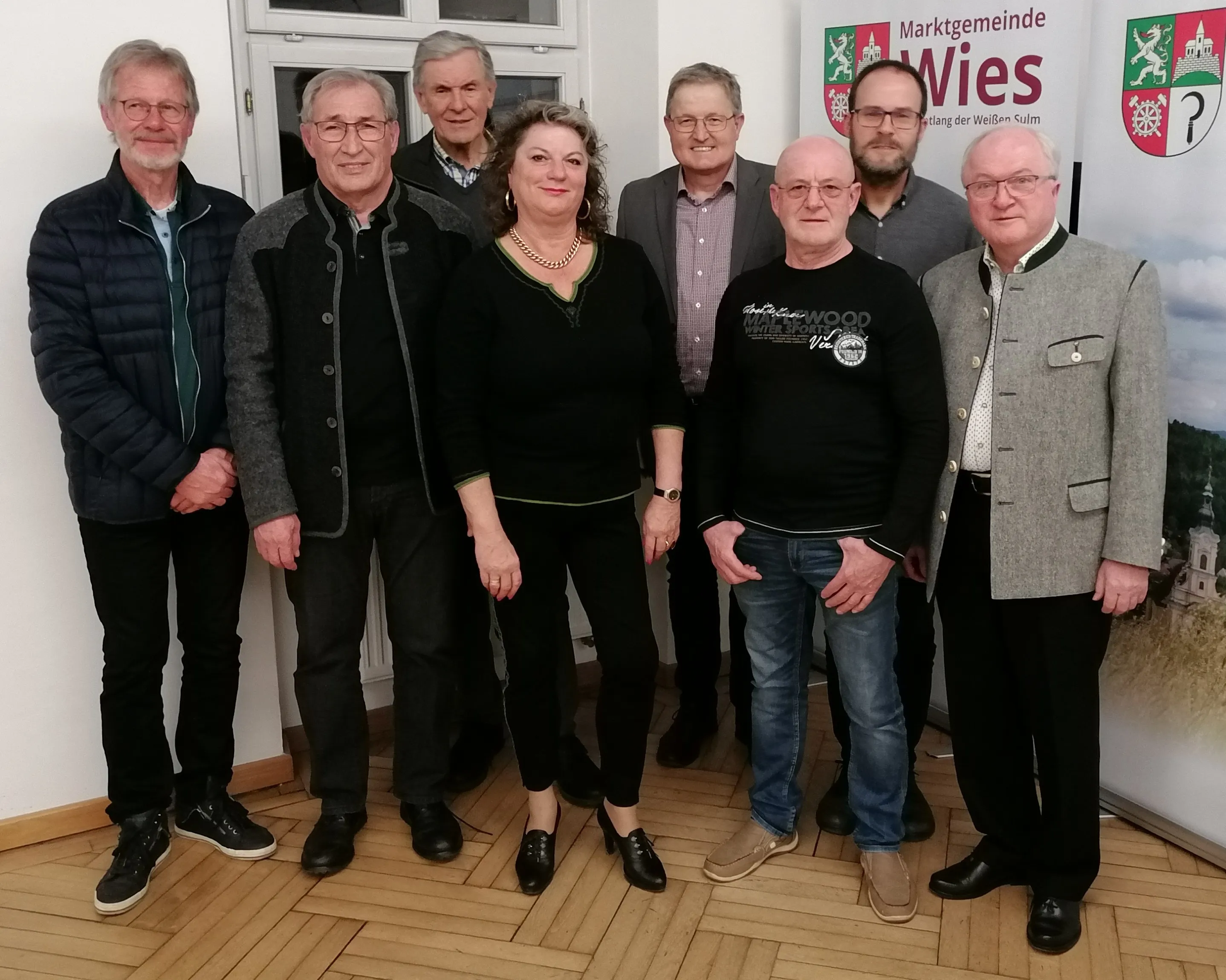 Sieben Erwachsene, Männer und Frauen, posieren für ein Gruppenfoto. Sie stehen vor einer weißen Wand mit einem Banner. Der Boden ist aus Holz.