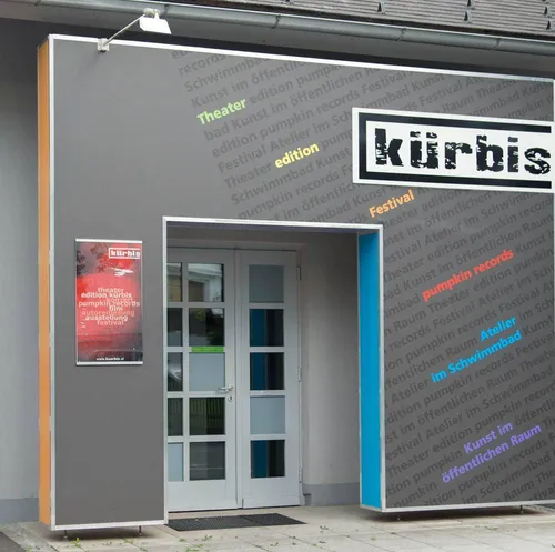 Eine graue Wand mit den Worten 'Theater', 'Edition' und 'Kürbis'. Eine Tür mit Glaspanelen und ein Schild für 'Kürbis Festival' darüber. Ein Plakat befindet sich auf der linken Seite der Tür.