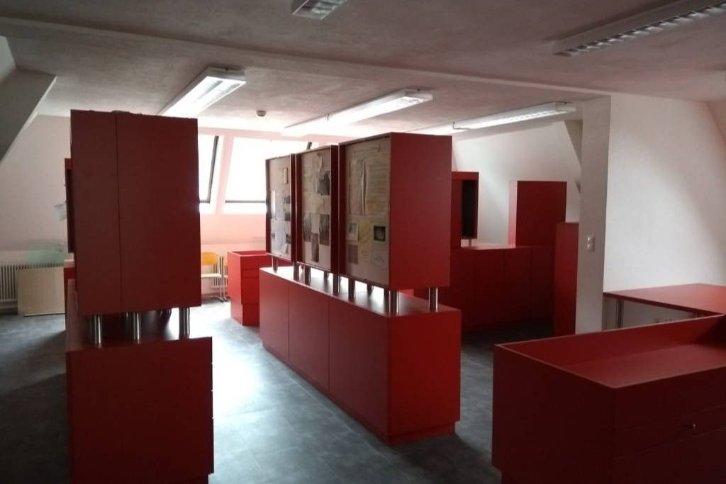 Ein leeres Büro mit roten Schränken und einer weißen Wand. Es gibt Leuchtstoffröhren an der Decke und Papiere auf den Schränken.