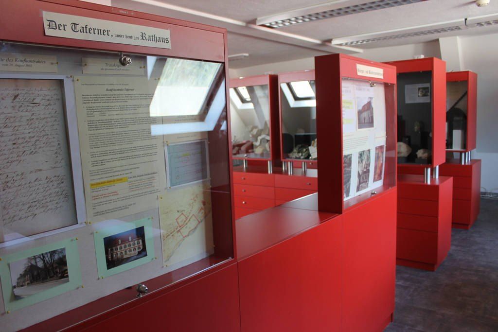 Ein Innenansicht eines Museums mit roten Vitrinen und einer Karte und Informationsplakaten auf der Glasplatte.