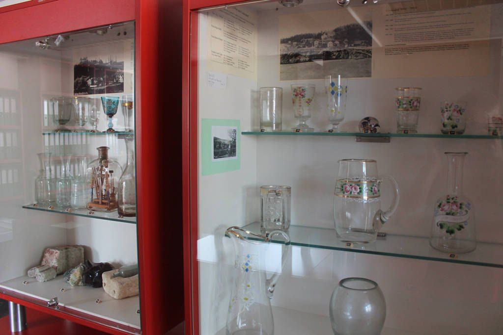 Ein Glasschrank mit roten Rahmen, der verschiedene Glaswaren und Glasartefakte präsentiert, mit dekorativen Elementen und Fotos an der Wand.