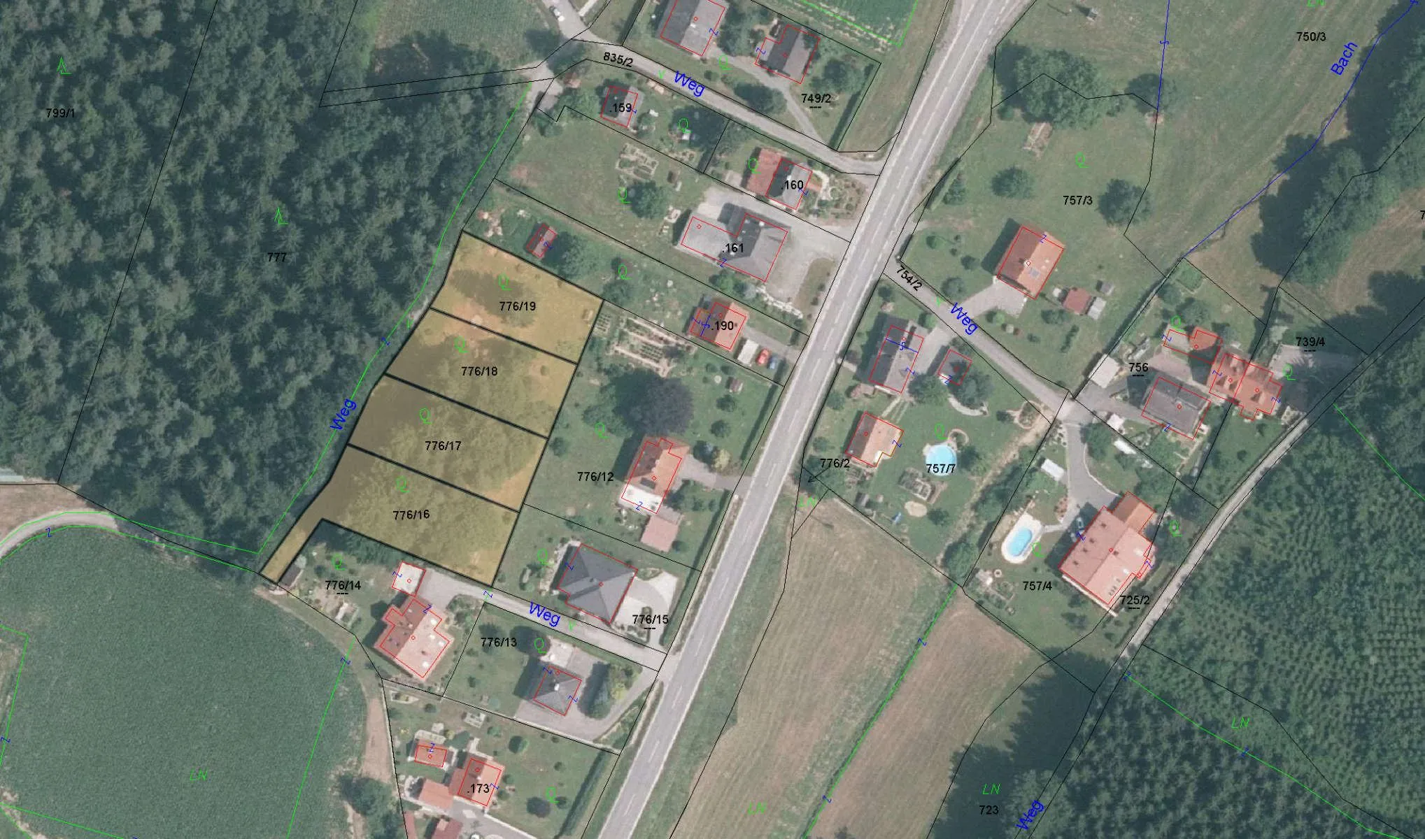 Luftaufnahme eines Wohngebiets mit mehreren Häusern und einer Straße. Jedes Haus hat eine nummerierte Bezeichnung und ist von einem eingezäunten Bereich umgeben. Bäume und Grünflächen sind um die Häuser herum vorhanden. Das Wohngebiet wird von einer Straße und einem Feld begrenzt.