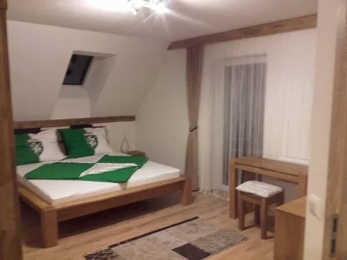 Ein gemütliches Schlafzimmer mit Holzboden, einem Bett, das mit grüner und weißer Bettwäsche geschmückt ist, einem Holzschreibtisch, einem Hocker und einem Fenster mit Vorhängen.