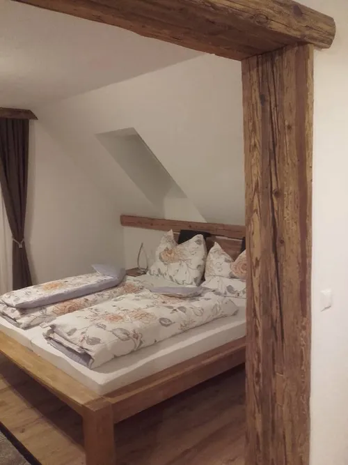 Ein gemütliches Schlafzimmer mit Holzbett, Blumenkissen und weißen Wänden. Ein Dachboden mit einem Holzbalken und einem Fenster ist sichtbar.