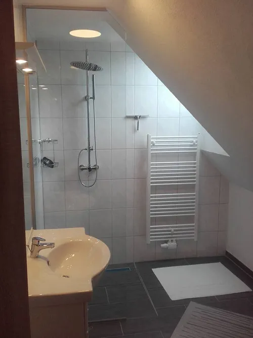 Ein kompaktes Badezimmer mit weißen Fliesen, einer Dusche, einem Waschbecken, einem Spiegel und einem Handtuchständer. Das Waschbecken hat einen Wasserhahn und einen Waschbeckenläufer.