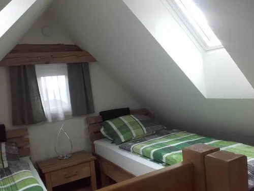 Ein Dachzimmer mit Holzmöbeln, einem mit grüner und weißer Bettdecke bedeckten Bett, einem Nachttisch mit Lampe und einem Fenster mit Vorhängen.