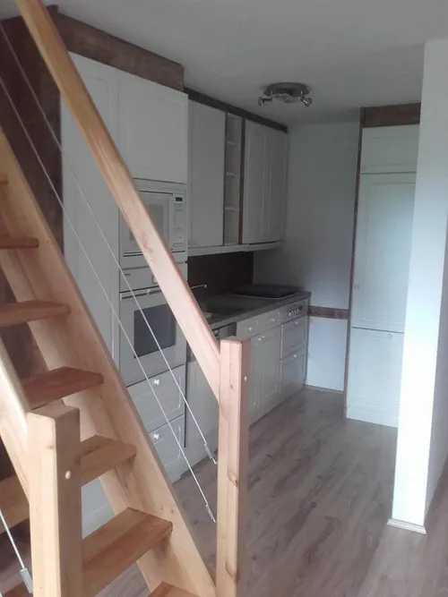 Eine kleine Küche mit Holztreppe, weißen Schränken, einer Mikrowelle, einem Ofen und einem Waschbecken. Der Raum hat Holzboden und eine Deckenlampe.