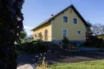 Ein helles gelbes Haus mit grauem Rand und mehreren Fenstern steht in einem ländlichen Gebiet. Es ist von einem Rasen mit Sträuchern und einer Einfahrt umgeben.