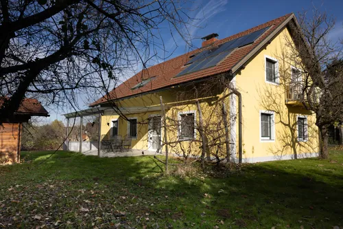 Ein gelbes Haus mit rotem Dach und Solarmodulen, umgeben von einem grünen Rasen, mit einer Terrasse mit Tisch und Stühlen und einem Baum vorne.