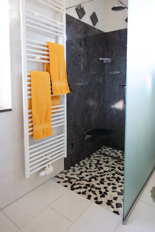 Ein modernes Badezimmer mit einem weißen Handtuchhalter, an dem drei gelbe Handtücher hängen. Der Duschbereich ist mit schwarzen und weißen Mosaiken gefliest. Der Duschkopf ist an einer Glaswand befestigt.