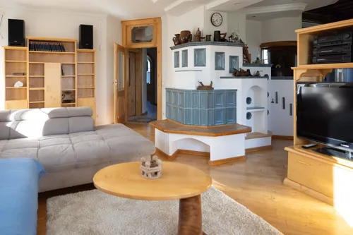 Ein gemütliches Wohnzimmer mit Holzboden und einem Kamin. Ein Sofa steht neben einem Holztisch mit einem dekorativen Gegenstand. Ein Bücherregal befindet sich links, und eine Tür führt in einen anderen Raum.
