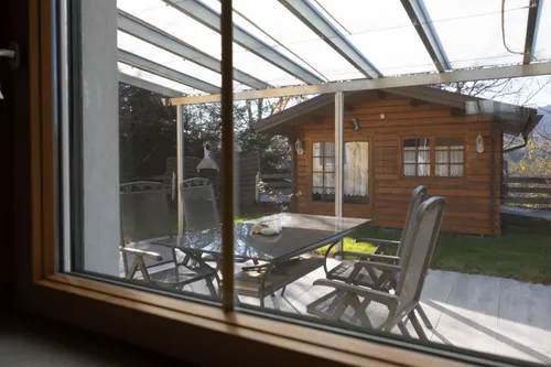 Eine Terrasse mit einem Glastisch und Stühlen unter einem Pergola. Im Hintergrund befindet sich ein Holzhaus mit Fenstern und einer Lampe.