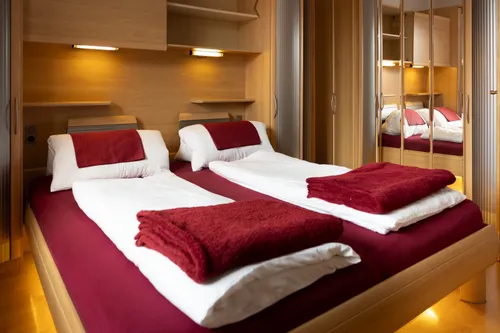 Zwei Einzelbetten mit roten Decken und Kissen, ordentlich gemacht, in einem Schlafzimmer mit Holzwänden und Regalen. Das Zimmer hat auch einen Schrank mit einem Spiegel.