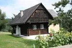 Ein kleines, rustikales Holzhaus mit grauem Dach und weißen Fenstern, umgeben von saftigem grünem Gras und Bäumen in einer ländlichen Umgebung.