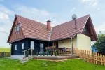 Ein kleines Haus mit rotem Dach und gelber Wand, auf einem Grasfeld gelegen. Vor dem Haus steht ein Holzzaun, und auf der Veranda befinden sich ein Tisch und Stühle.