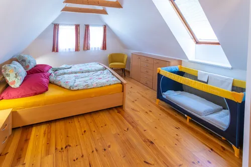Ein Dachgeschosszimmer mit Holzboden, einem Bett mit Blumenbezug, einem gelben Stuhl, einem Kinderbett und Holzschränken. Das Zimmer hat zwei Fenster mit Vorhängen.