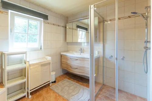 Ein modernes Badezimmer mit einer Glasdusche, einem Waschbecken und einem Schrank. Ein Fenster mit Vorhängen sorgt für natürliches Licht. Ein Teppich liegt auf dem Boden und ein kleiner Mülleimer ist in der Nähe.