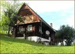 Ein Holzhaus mit Balkon und Schornstein liegt auf einem grünen Hügel, umgeben von üppigem Baumbestand und klarem Himmel.
