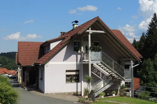 Ein weißes Haus mit rotem Dach und Glastreppe. In der Nähe der Treppe und Fenster befinden sich Topfpflanzen. Ein Schornstein ist auf dem Dach. Die Nummer 117 steht an der Wand.