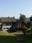 Ein Haus mit einem grauen Dach und weißen Wänden steht auf einem grasbewachsenen Hügel. Es ist von üppigen grünen Bäumen und Pflanzen umgeben. In der Ferne befinden sich Hügel unter einem klaren blauen Himmel.