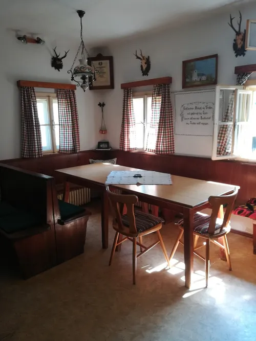 Eine gemütliche Essecke mit Holzbänken, einem Holztisch und Stühlen. Zwei Fenster mit Vorhängen. Eine Kronleuchter und Bilderrahmen schmücken die Wände.