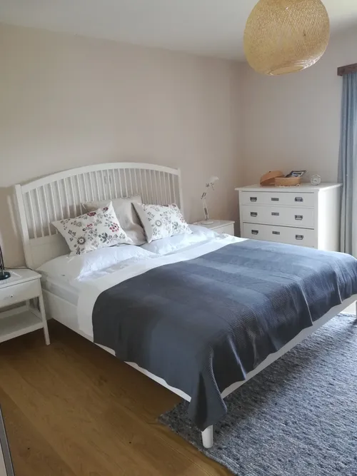 Ein Schlafzimmer mit einem weißen Bett, einem weißen Kopfteil und einer blauen Decke. Zwei Nachttische befinden sich auf beiden Seiten, mit einer Lampe auf der rechten Seite. Ein weißer Anzugskasten steht an der Wand, mit Büchern und einer Uhr oben.