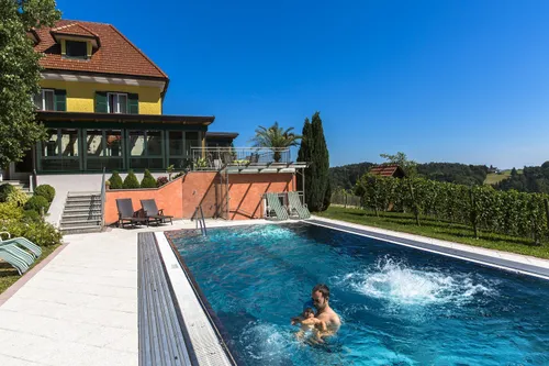 Ein Mann und ein Kind schwimmen in einem großen Außenpool. Der Pool ist von einer Terrasse mit Liegestühlen umgeben. Die Umgebung ist eine Villa mit einer Terrasse und einem Garten. Der Himmel ist klar und blau.