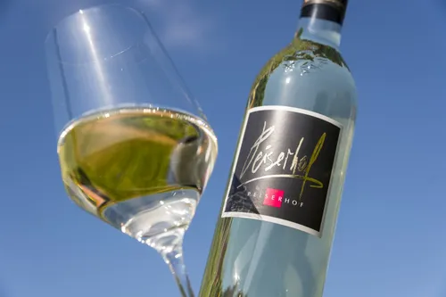 Eine Flasche Weißwein, mit dem Etikett 'Peiserhof', neben einem kristallklaren Glas Wein unter blauem Himmel.