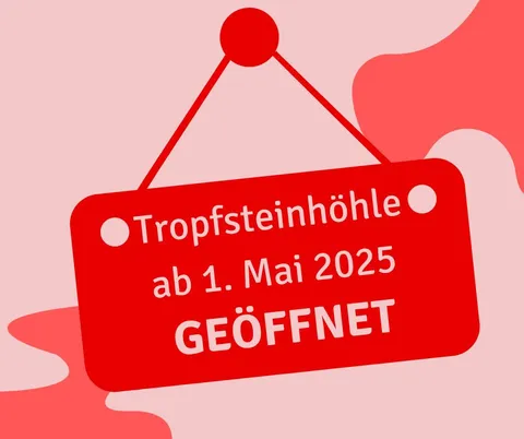 Ein rotes Schild mit weißem Text steht 'Tropfsteinhöhle ab 1. Mai 2025 GEÖFFNET'. Das Schild hängt an einem roten Faden vor einem rosa Hintergrund.