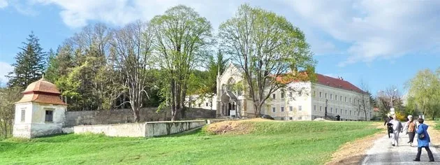 Ein historisches Gebäude mit rotem Dach und Rundbogenfenstern steht in einem üppigen grünen Park, an dem Menschen vorbeigehen.